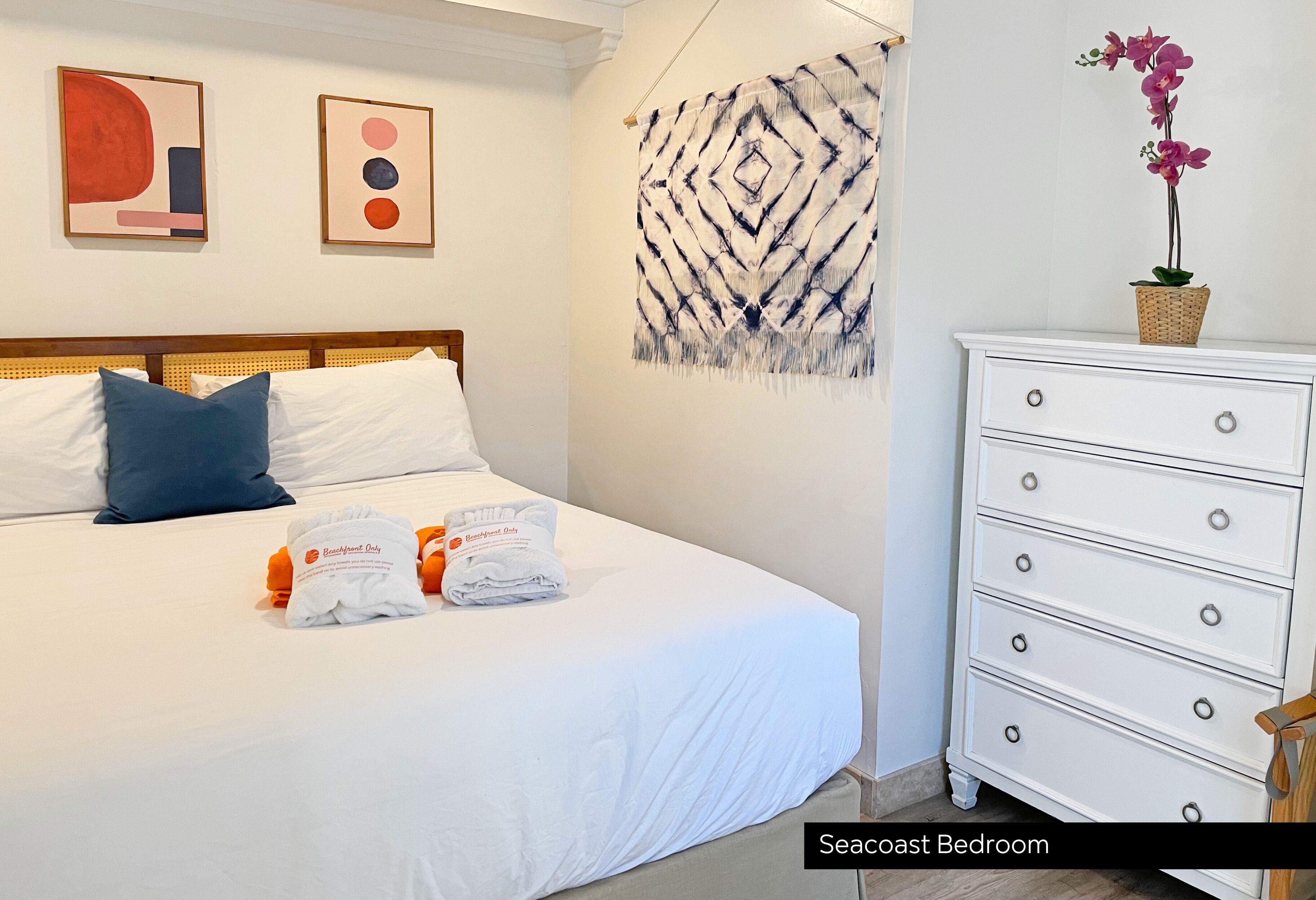 Un dormitorio en la costa con una cama bien hecha, un aparador blanco y un tapiz de pared.