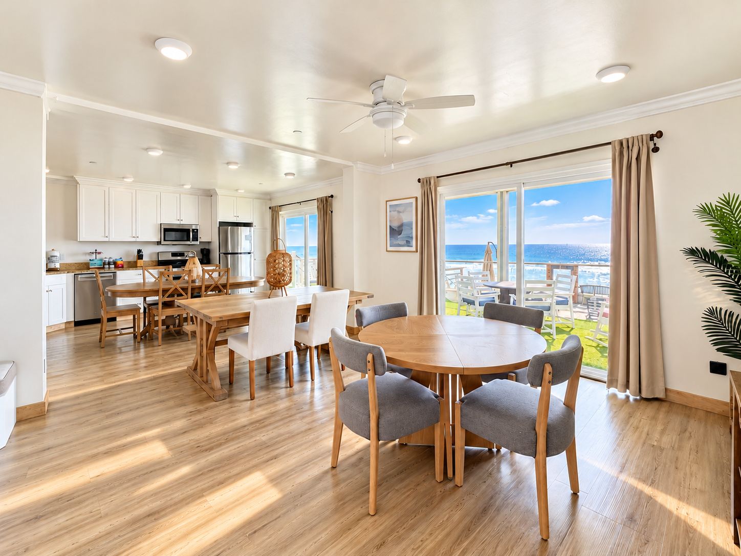 A spacious living area with a large round table and chairs, a kitchen with white cabinets, and a balcony with a view of the ocean.