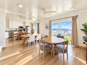 A spacious living area with a large round table and chairs, a kitchen with white cabinets, and a balcony with a view of the ocean. - Sand, Sun, Serenity P1111-23 (Oceanside)