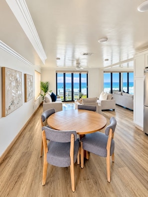 A spacious living area with a round wooden table surrounded by chairs, a sofa set, and a view of the ocean. - Sand, Sun, Serenity P1111-23 (Oceanside)