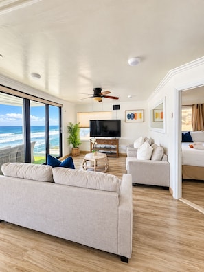 A spacious living area with a large window overlooking the ocean, a ceiling fan, a flat-screen TV, and a seating arrangement with sofas and a coffee table. - Sand, Sun, Serenity P1111-23 (Oceanside)
