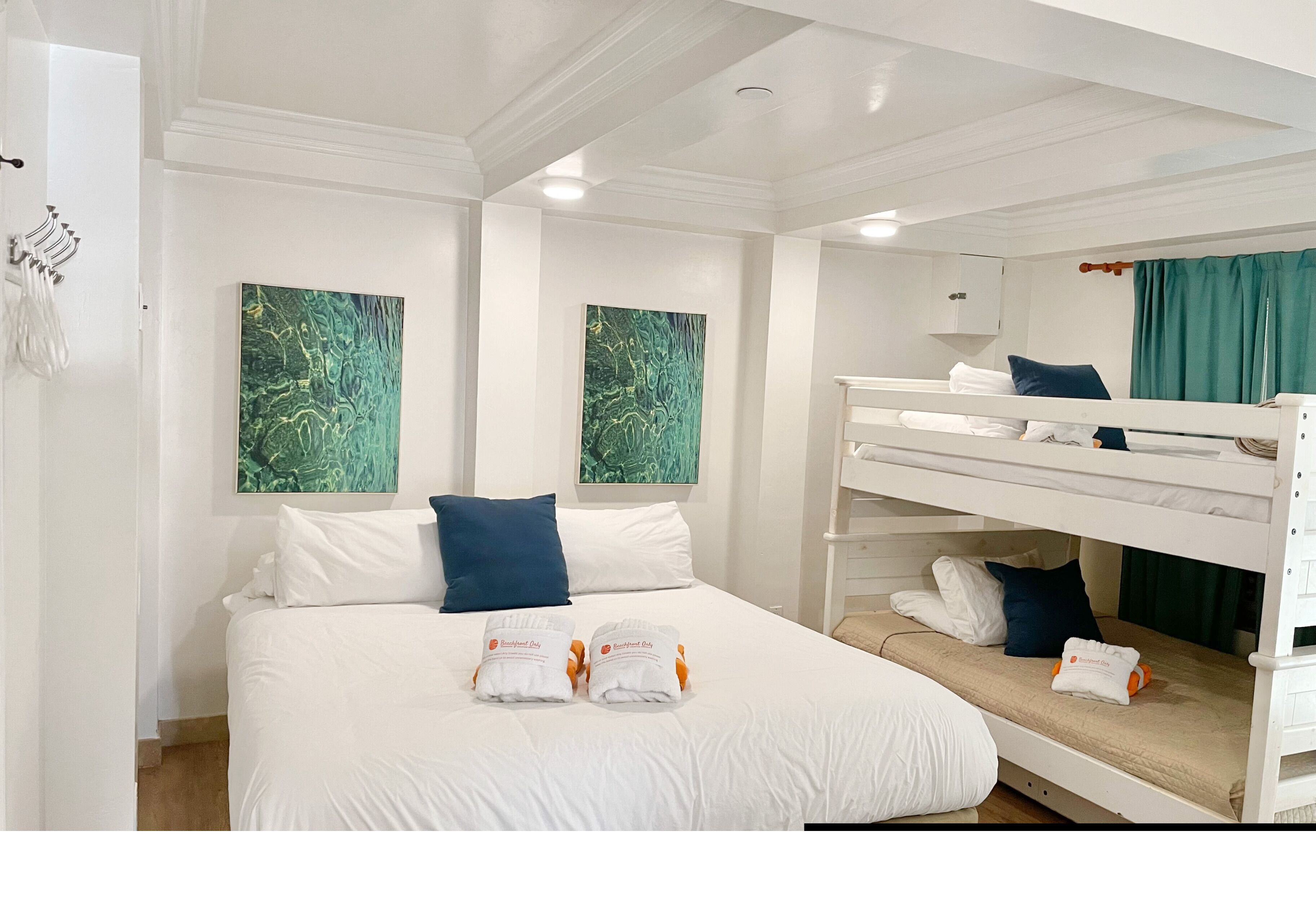 Une chambre d’hôtel avec un lit superposé, un grand lit avec du linge de lit blanc, deux tableaux abstraits verts accrochés aux murs et une armoire blanche.