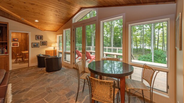 A cozy room with a stone floor, wooden ceiling, and large windows offering a view of trees. There is a round table with chairs and a small seating area with a bench and chairs. - Luxury Lake Escape in Quiet Cove w/ Dock (Oakland)