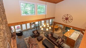 A spacious loft-style living area with a high ceiling, large windows, a central chandelier, and a stone fireplace. - Luxury Lake Escape in Quiet Cove w/ Dock (Oakland)