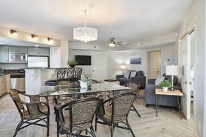 A modern kitchen with built-in appliances, a dining area with wicker chairs, a ceiling fan, and a living room with a sofa and lamp. - Oceanview Retreat, 302 (Tybee Island)
