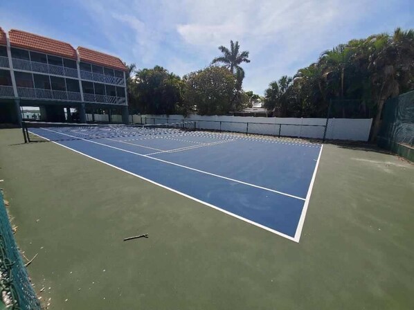 Een tennisbaan met net, omringd door een hek en palmbomen.