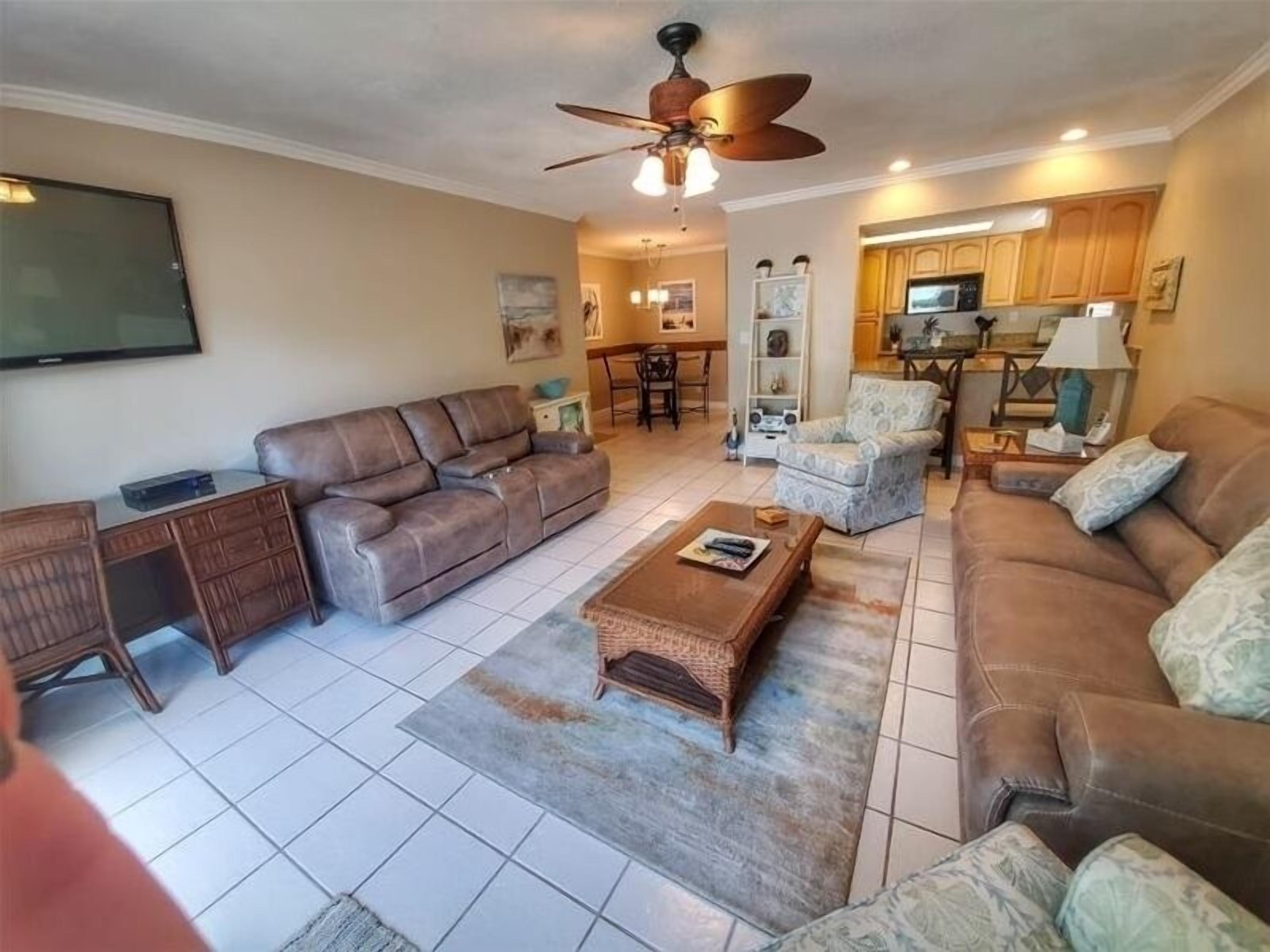 A living room with a brown leather sofa, a coffee table, and a patterned rug. There is a ceiling fan, a television mounted on the wall, and a kitchen area in the background.