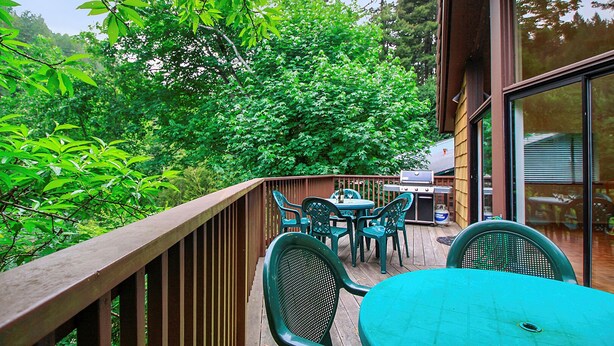 An outdoor deck with a wooden railing, a barbecue grill, and green plastic chairs and tables. - Eric's Landing ~ Riverfront, Fun with Friends Getaway! Dog Friendly! (Guerneville)