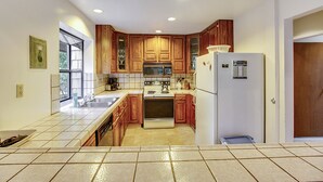 A kitchen with wooden cabinets, a white refrigerator, and a tiled floor. - Eric's Landing ~ Riverfront, Fun with Friends Getaway! Dog Friendly! (Guerneville)