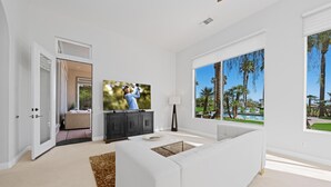 Una sala de estar moderna con un sofá blanco, un televisor de pantalla plana, un mueble de madera oscura y un ventanal con vistas a palmeras y una piscina.