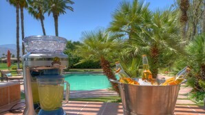 Ambiente junto a la piscina con una licuadora y un balde de botellas, rodeado de palmeras y una piscina cristalina.