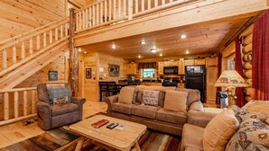 A log cabin interior with a staircase, a living room with a sofa, a coffee table, and a kitchen area.