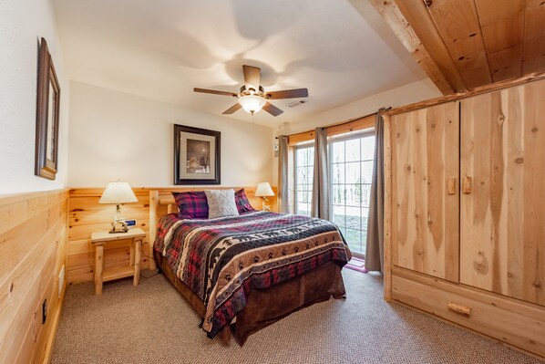A cozy bedroom with a wooden ceiling fan, a bed with a patterned blanket, two bedside tables with lamps, and a large wooden wardrobe.