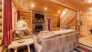 A log cabin interior with a fireplace, a sofa, a table with a lamp, and a staircase.
