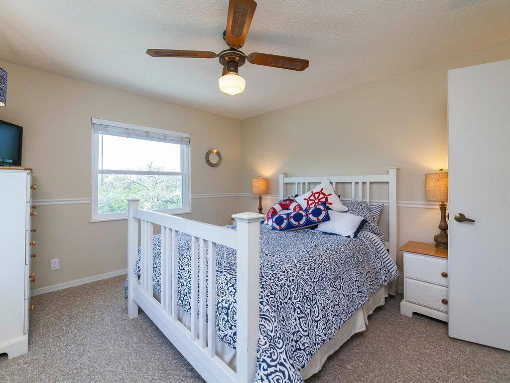 A bedroom with a white crib, a bed with patterned bedding, a ceiling fan, a window, and a nightstand with a lamp.