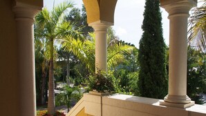 Un espacio al aire libre cubierto con columnas, una escalera de ladrillo y abundante vegetación, incluyendo palmeras y plantas con flores.