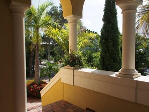 A covered outdoor area with columns, a brick staircase, and lush greenery including palm trees and flowering plants.