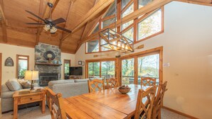 A spacious living area with a wooden dining table, comfortable seating, a stone fireplace, and large windows offering views of the outdoors. - Spacious Cedar Chalet w/ Hot Tub & Pool Table (Swanton)