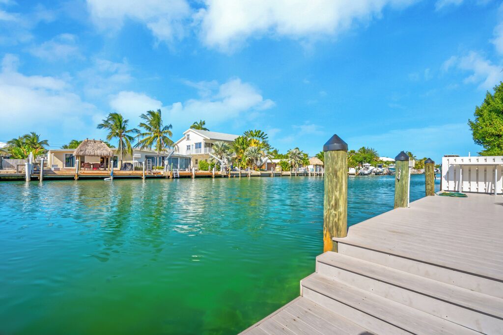 A wooden dock extending into a clear blue water body with houses and palm trees in the background.
