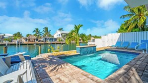 Un área de piscina con sillas de descanso, vista al agua y palmeras.