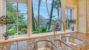 A kitchen with a large window overlooking a tropical garden and the ocean. It features a granite countertop, stainless steel sinks, and white cabinetry. - P05 - 5 bedroom Ocean Front home with Private pool and deep water dockage. (Key Colony Beach)