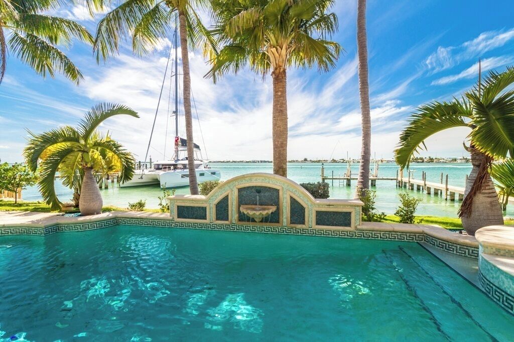 A swimming pool with a decorative edge, surrounded by palm trees and a view of the ocean with boats.