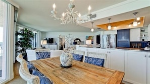A spacious dining area with a wooden table and white upholstered chairs, a decorative vase centerpiece, and a modern kitchen in the background.
