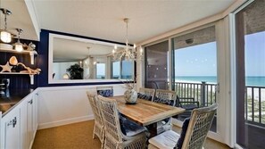Ein Essbereich mit Holztisch, Korbstühlen, einer Deckenleuchte und einem großen Fenster mit Blick auf den Strand.