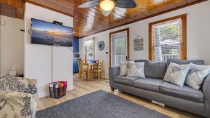 A cozy living room with a grey sofa, a television mounted on the wall, a ceiling fan, and a dining area with a table and chairs. - Reef at The Arbors—Charming 50s Cottage Steps from Beach w/ Pool & Lush Grounds (Indian Rocks Beach)
