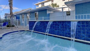 A swimming pool with a waterfall feature, surrounded by a blue-tiled wall and a white fence. - Reef at The Arbors—Charming 50s Cottage Steps from Beach w/ Pool & Lush Grounds (Indian Rocks Beach)
