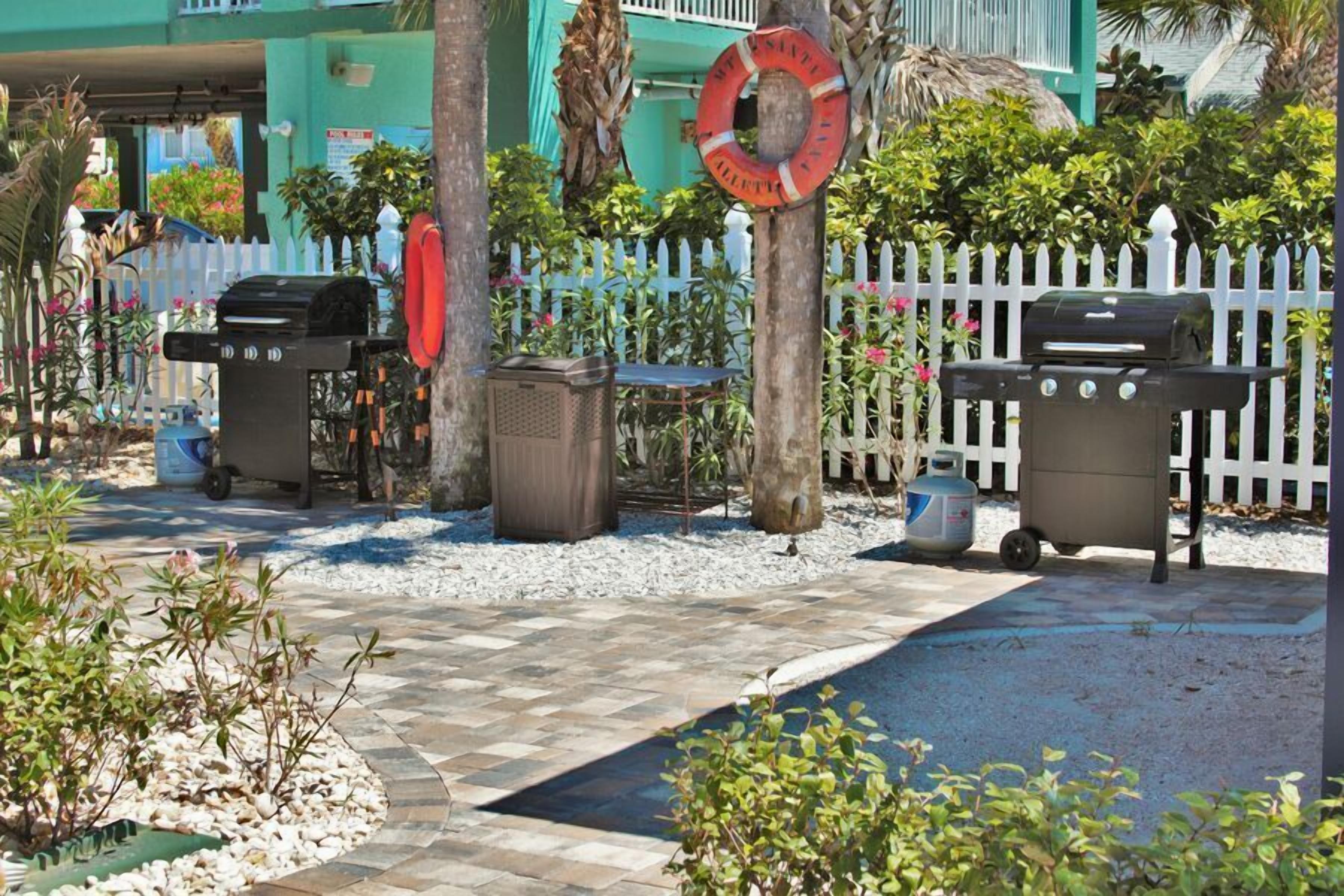 Un patio exterior con dos parrillas de barbacoa, una valla de piquetes blancos y un flotador de piscina colgado de un árbol.