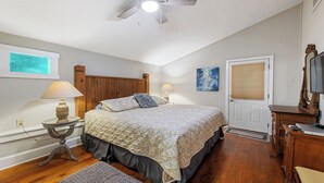 Une chambre à coucher avec une tête de lit en bois, un lit recouvert d’une courtepointe, deux lampes de chevet, un tableau, une porte avec une fenêtre et une commode en bois dotée d’un miroir.
