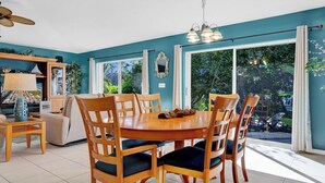 A dining area with a round table and chairs, a ceiling fan, and a view of greenery outside.