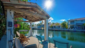 A wooden deck with lounge chairs and a covered area overlooking a calm body of water, with buildings in the background.