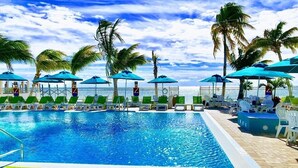 A swimming pool with lounge chairs and umbrellas, surrounded by palm trees and a view of the ocean.