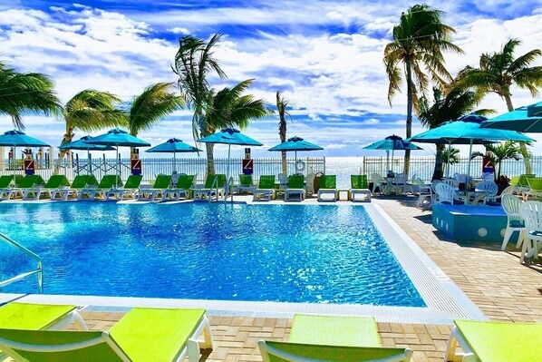A swimming pool with lounge chairs and umbrellas, surrounded by palm trees and a view of the ocean.