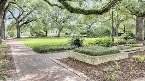 A pathway flanked by large trees with moss-covered branches, leading to a grassy area with a building in the background. - Congress Garden Suite- Off Street Parking Included! (Savannah)