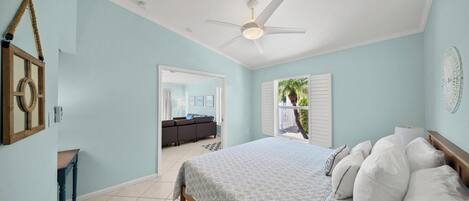 A bedroom with a bed, a ceiling fan, a view of a living area, and a door leading outside.