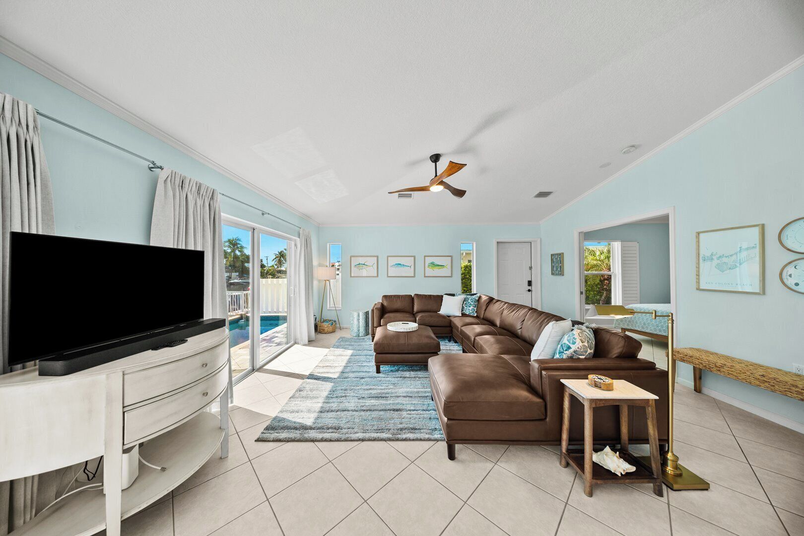 A living room with a flat-screen TV, a brown leather sofa, a ceiling fan, and a view of a pool.