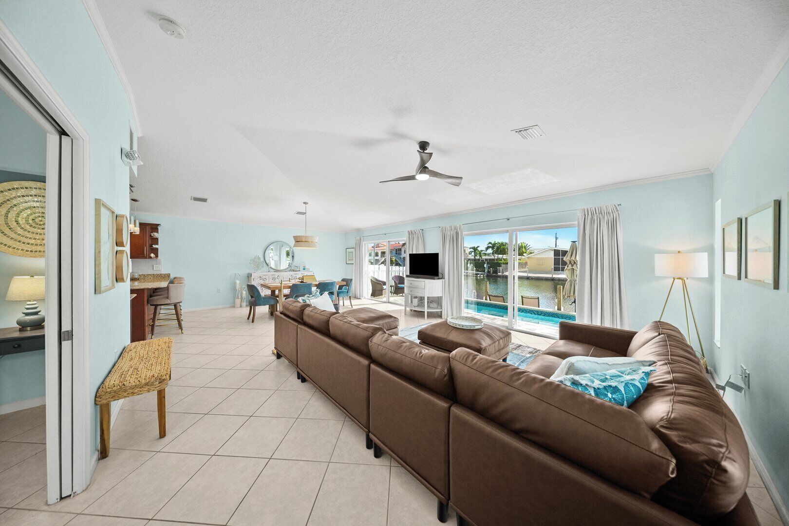 A spacious living room with a large brown sofa, a dining area with a table and chairs, a TV, and a view of a swimming pool.