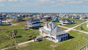 An aerial view of a coastal residential area with houses featuring balconies, a prominent white building with a grey roof, and a clear view of the ocean in the background. - A Sea Glass Estate: 9  BR, 5  BA House in Crystal Beach, Sleeps 34 (Bolivar Peninsula)