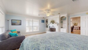 A bedroom with a large bed, a gray sofa, a fireplace, and a dining area visible through an open door. - A Sea Glass Estate: 9  BR, 5  BA House in Crystal Beach, Sleeps 34 (Bolivar Peninsula)