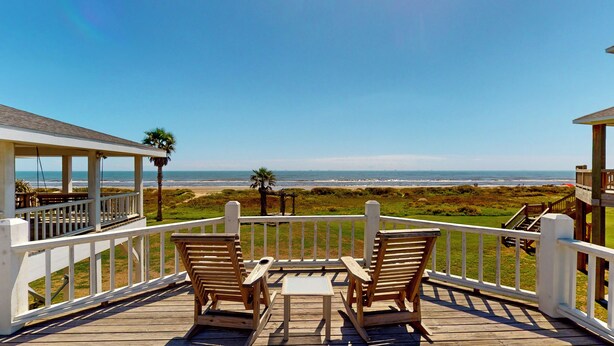 A wooden deck with two chairs and a table, overlooking a beach with a clear view of the ocean. - A Sea Glass Estate: 9  BR, 5  BA House in Crystal Beach, Sleeps 34 (Bolivar Peninsula)