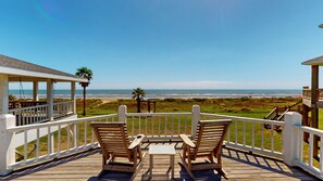 A wooden deck with two chairs and a table, overlooking a beach with a clear view of the ocean. - A Sea Glass Estate: 9  BR, 5  BA House in Crystal Beach, Sleeps 34 (Bolivar Peninsula)