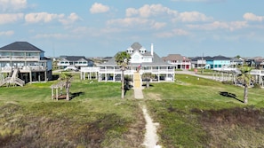 Een woongebied aan de kust met meerdere huizen, waaronder een centraal gelegen wit huis met veranda, en een zandpad dat er naartoe leidt.