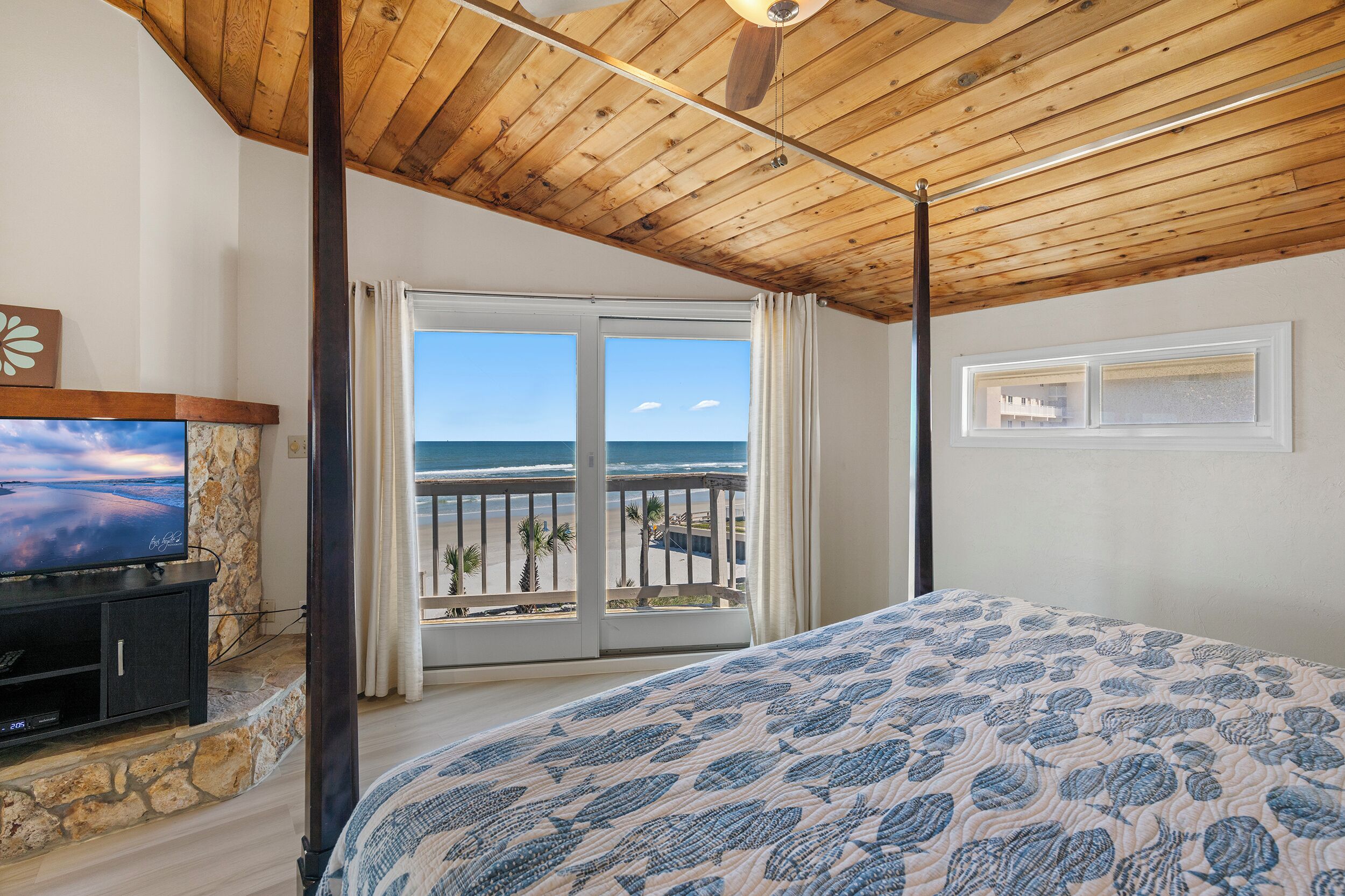 Ein Schlafzimmer mit einem großen Bett, einem Fernseher auf einem Steinkamin und einem Balkon mit Blick auf den Strand.
