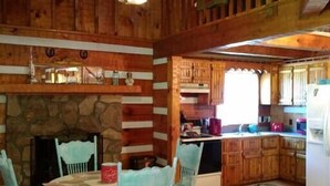 A rustic cabin interior with a stone fireplace, wooden dining table, and chairs, a kitchen with wooden cabinets, and a ceiling fan.