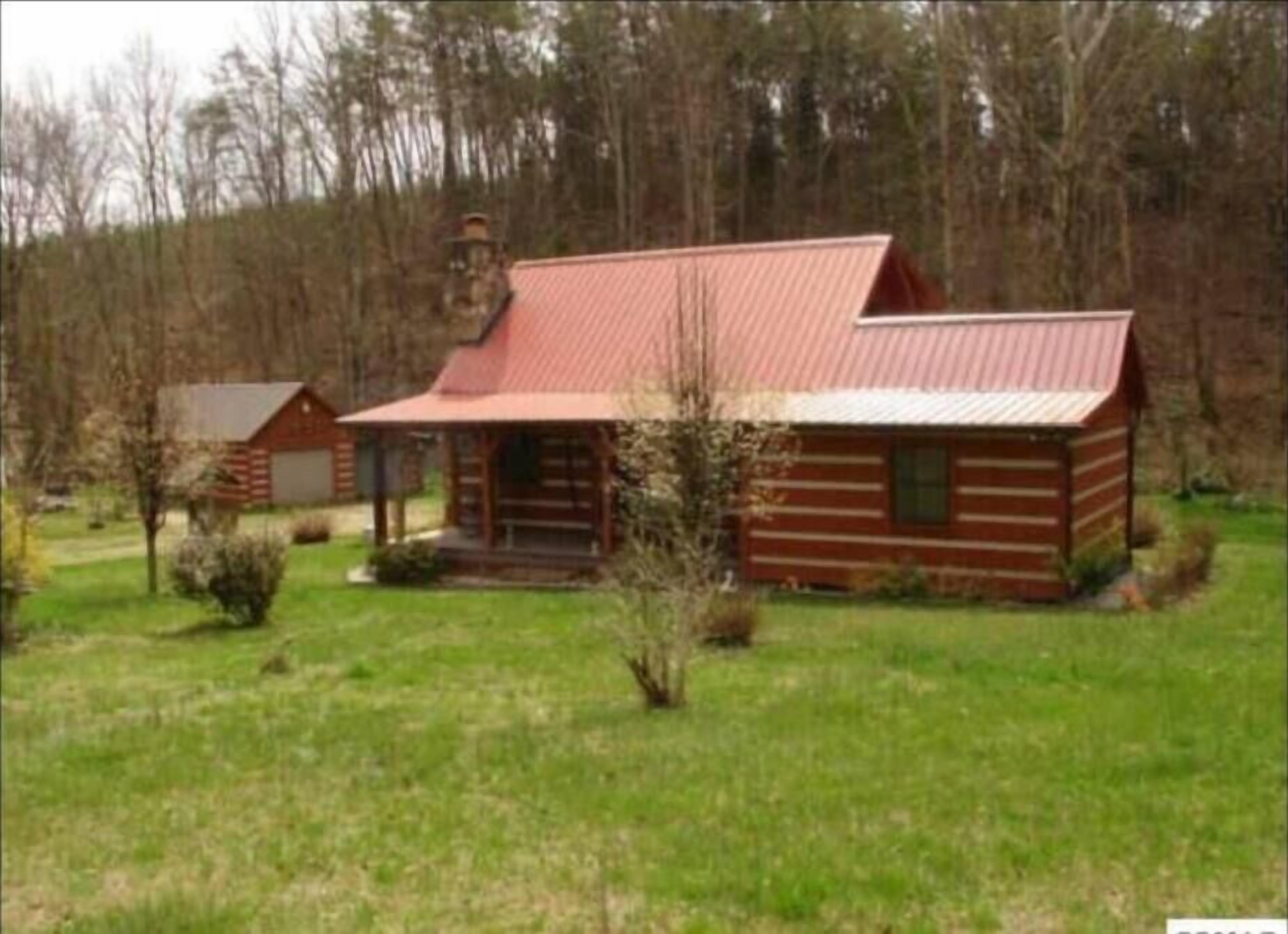 Une cabane rustique en rondins de bois, surmontée d’un toit en métal, entourée d’une cour verdoyante et d’arbres.