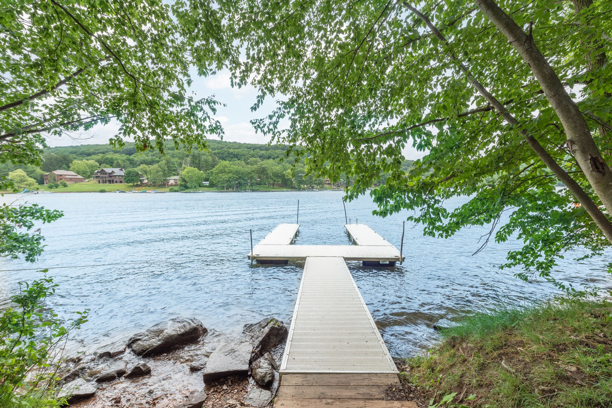 Eine Holzsteganlage erstreckt sich über einen stillen See, umgeben von üppiger Vegetation und Gebäuden in der Ferne.