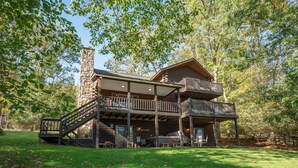 A two-story log cabin with a stone chimney, wooden balconies, and a covered porch area. - Enjoy Adventure & Relaxation w/ Hot Tub & Fire Pit (McHenry)